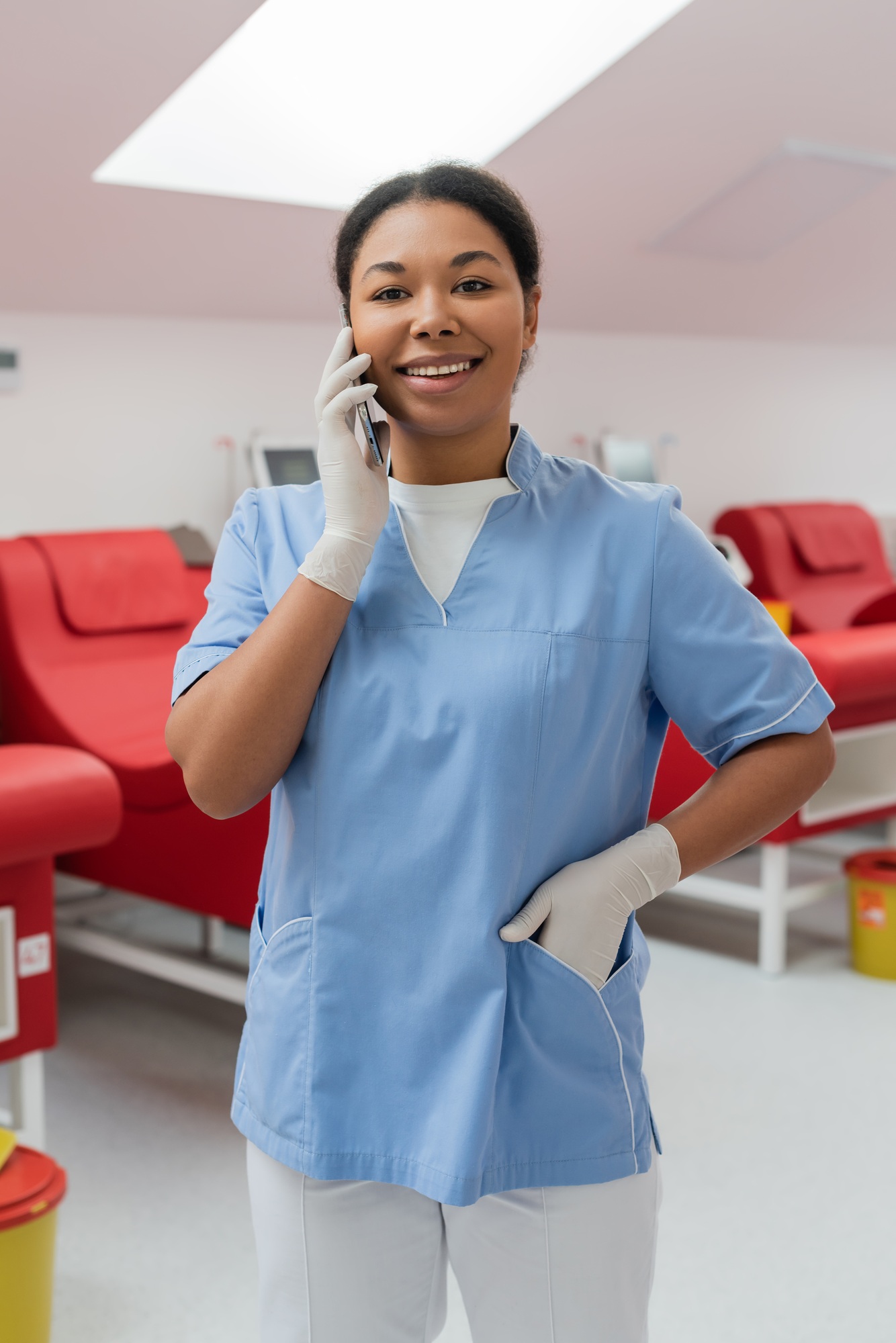 joyful multiracial nurse in latex gloves standing with hand in pocket of blue uniform and talking